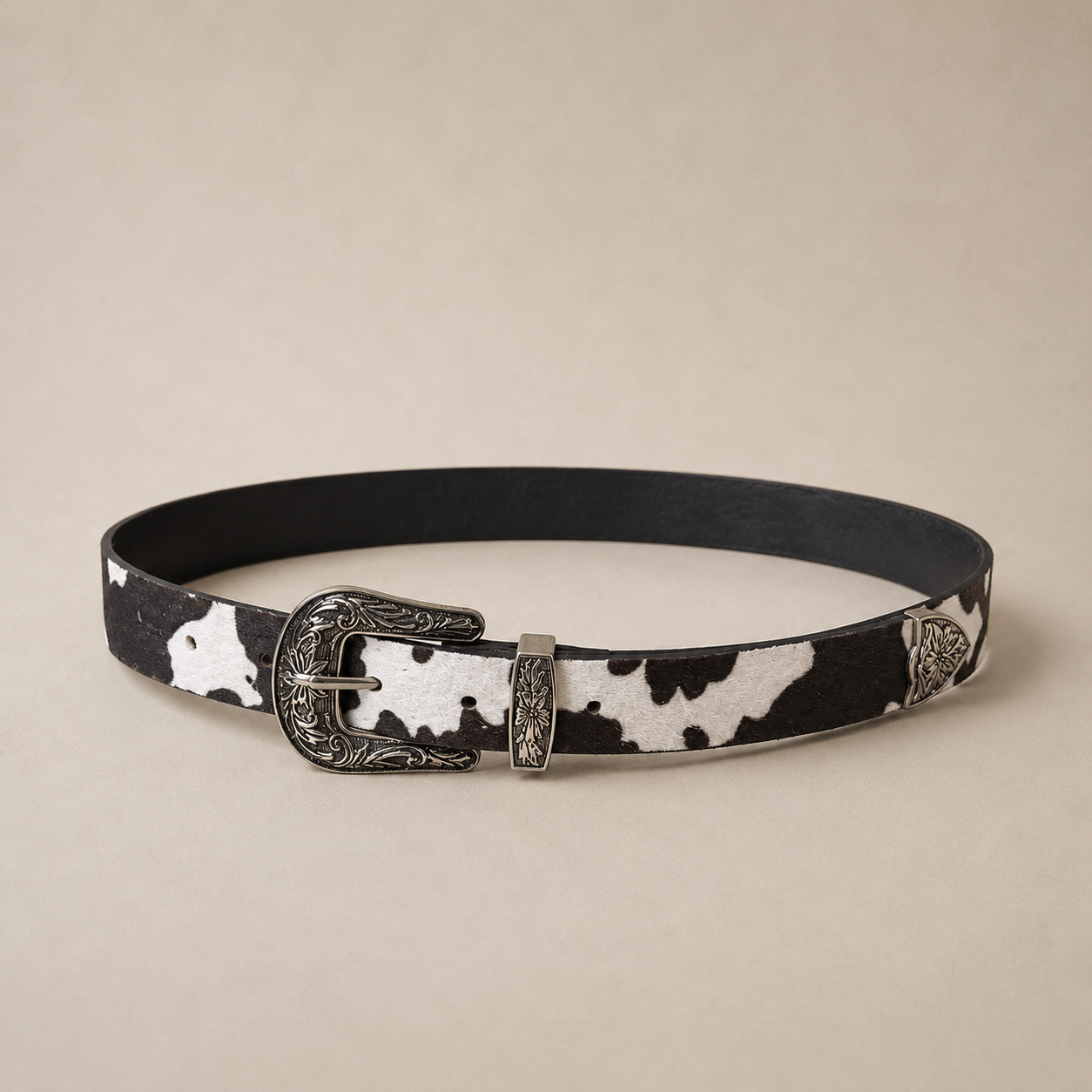 Cinto fino con estampado cow print blanco y negro y hebilla metálica plateada de estilo western con detalles grabados.