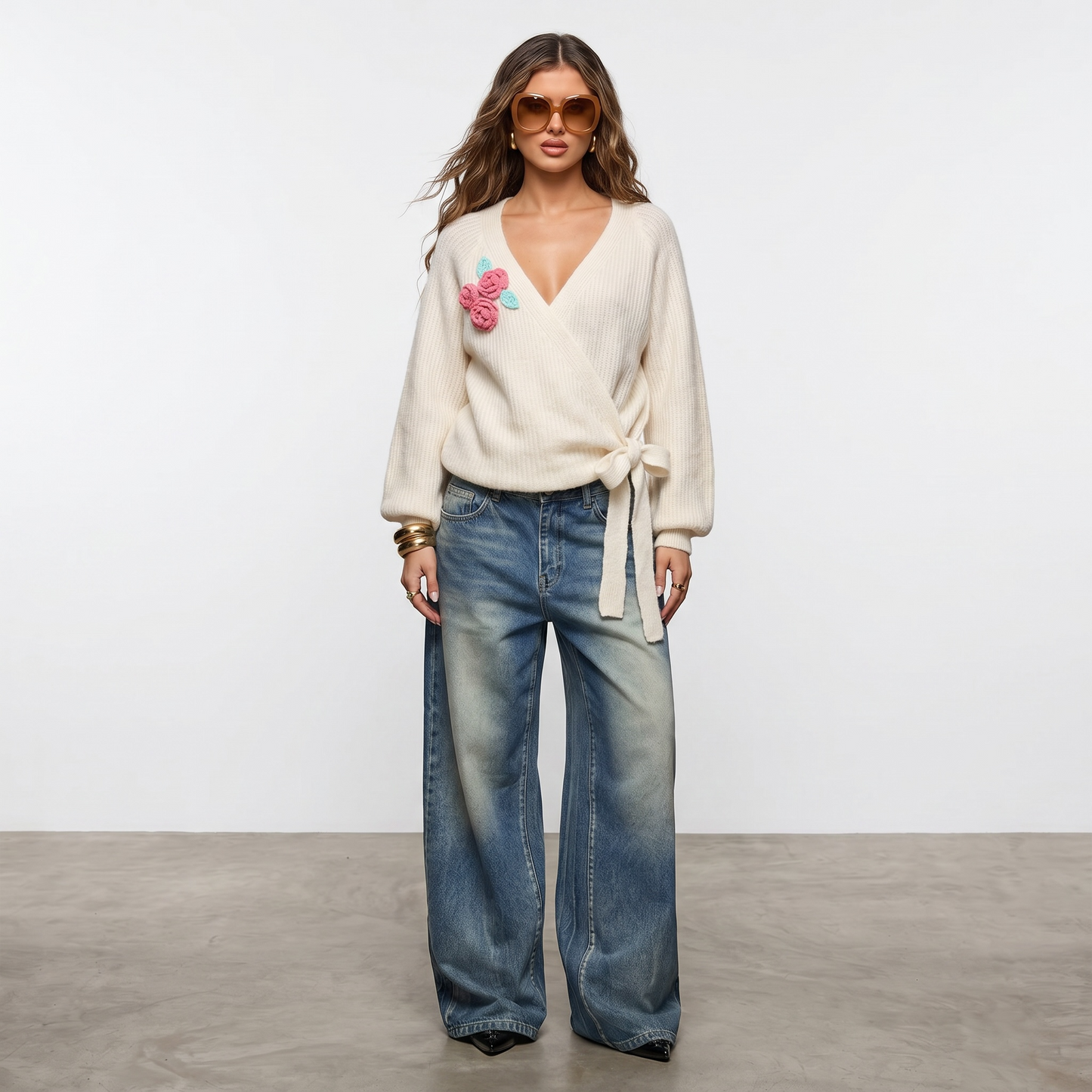 Mujer luciendo sweater tejido cruzado color crudo con flores aplicadas en relieve y lazo lateral, combinado con jean amplio de estilo urbano. Prenda excusiva Paola Santos